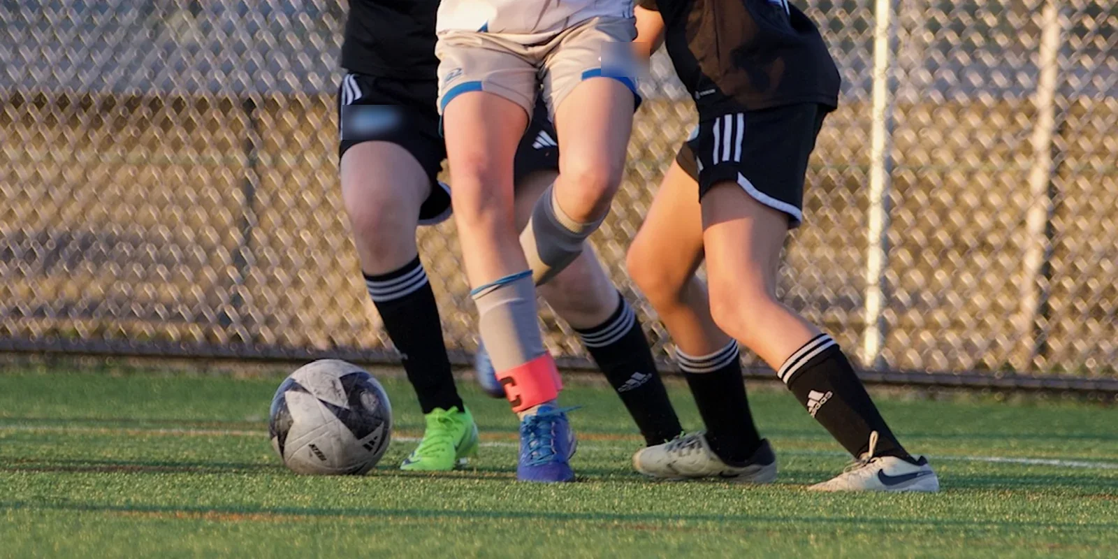 Four youth players close on a loose ball on artificial turf at golden hour, shown from the knees down.